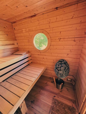 A sauna room with wooden benches, a circular window, a stone heater, and a metal container labeled 'HUM'.