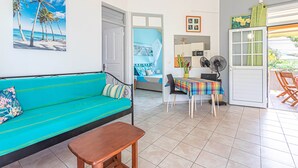 A room with a teal sofa, a wooden table, a dining area with a colorful tablecloth, a fan, and a view of the outdoors through a door. - ALAMANDA (1 to 3 people) (DESHAIES)