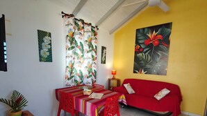 A living room with a red sofa, a dining table with red chairs, a potted plant, and tropical-themed artwork on the walls. - PAPYRUS (1 to 3 people) (DESHAIES)
