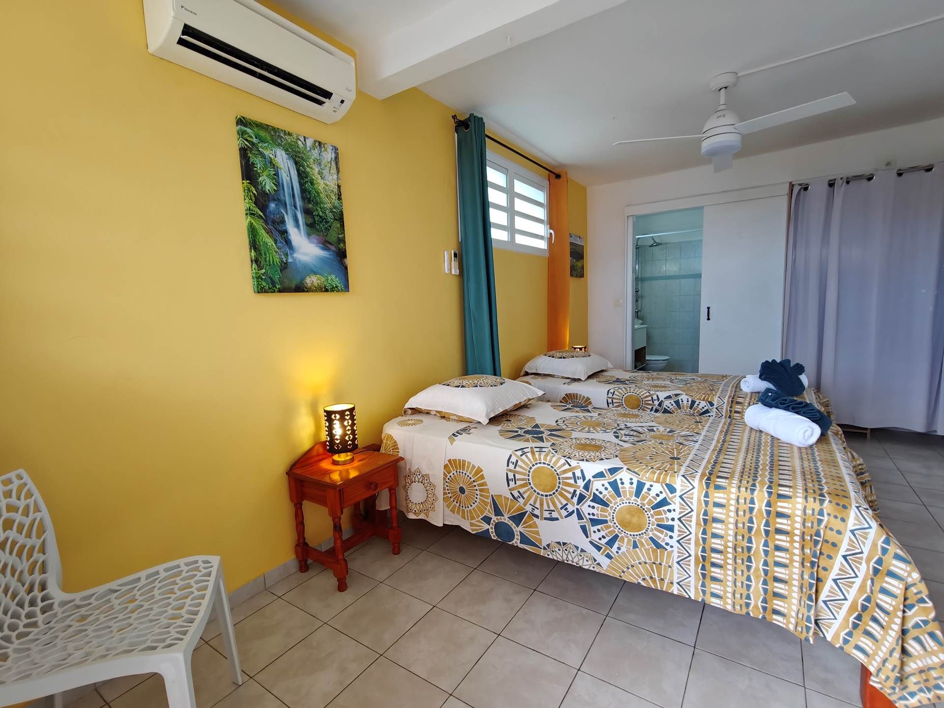 Habitación de hotel con cama, mesita de noche, una silla, un cuadro y baño visible a través de una puerta abierta.