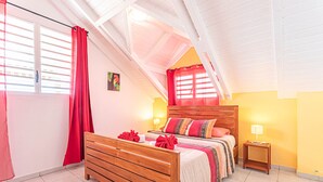 Slaapkamer met een houten bed, rode gordijnen, een klein nachtkastje met een lamp en een raam met jaloezieën.