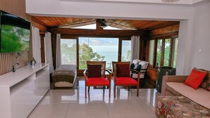 A spacious living area with a large window offering a view of the sea, a flat-screen TV mounted on the wall, and a sofa with a floral cover. - Casa Praia das Cigarras (Sao Sebastiao)