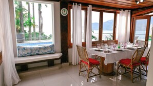 A dining area with a table set for four, wooden chairs with red cushions, a bench with a blue cushion, and a view of palm trees and mountains through large windows. - Casa Praia das Cigarras (Sao Sebastiao)
