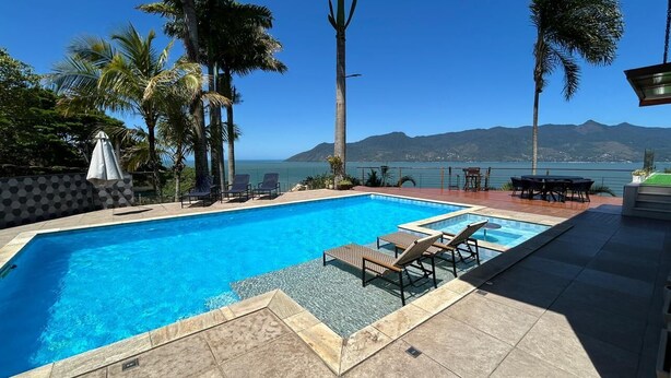 A rectangular swimming pool with lounge chairs and a table, surrounded by palm trees and a view of the sea and mountains. - Casa Praia das Cigarras (Sao Sebastiao)