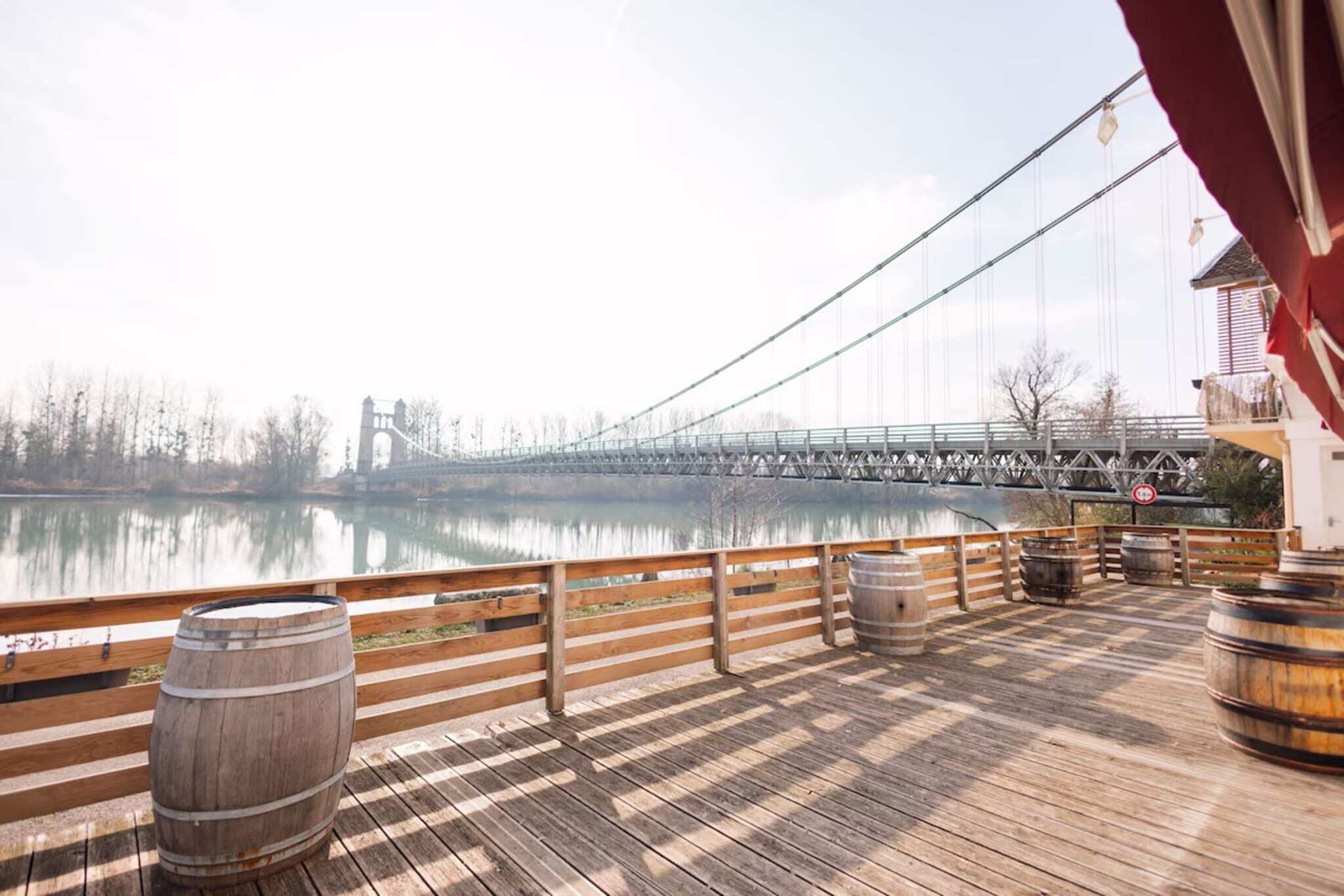 A wooden deck with barrels, overlooking a calm river and a bridge.