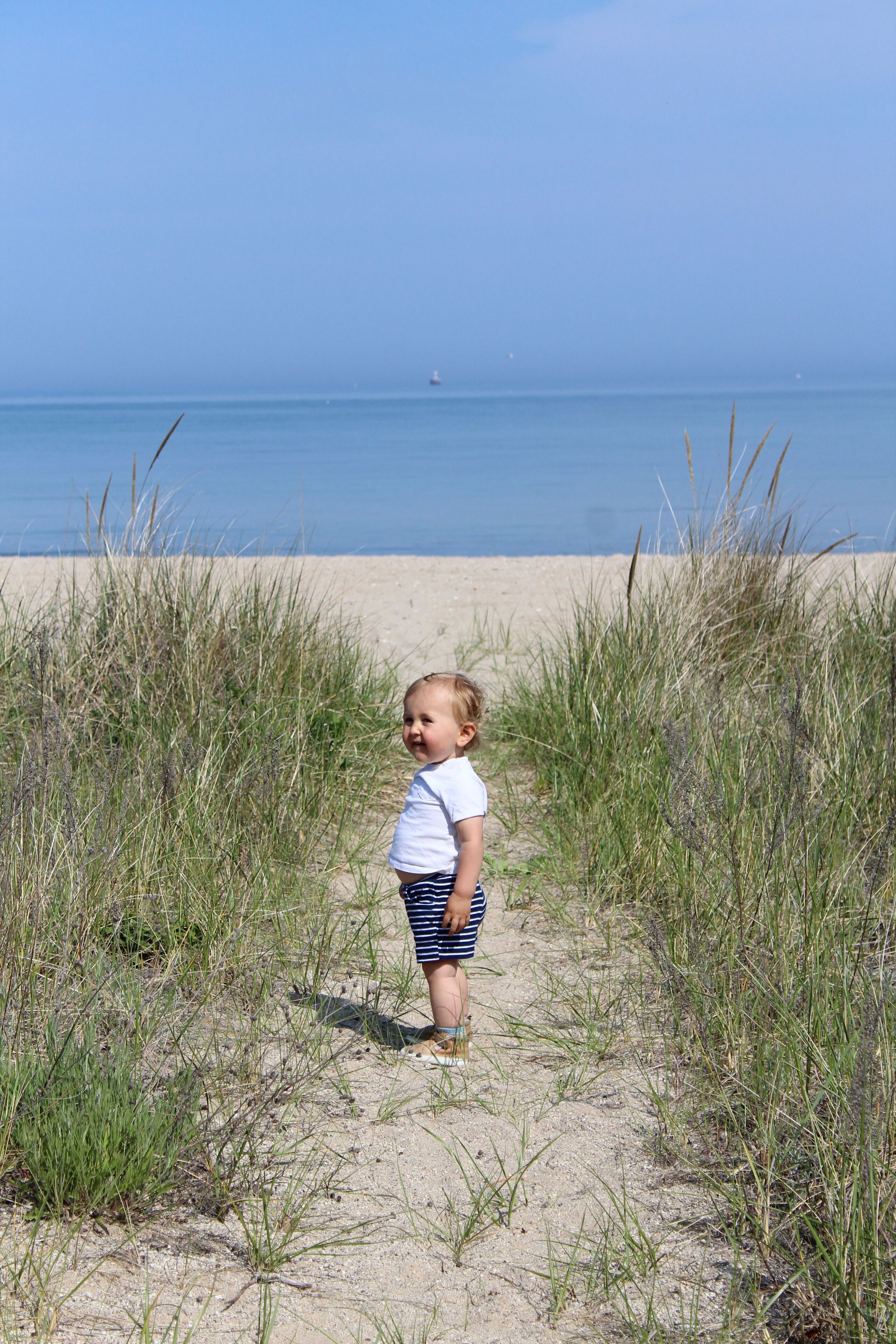 A child standing on a sandy beach with tall grass in the foreground and a calm sea in the background.