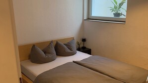 Ein Einzelbett mit Holz-Kopfteil, zwei Kissen, ein Nachttisch mit Lampe und ein Fenster mit Pflanze.