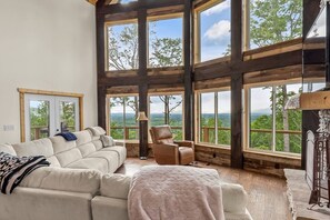 A spacious room with a large window, a sofa, a chair, and a lamp. - Mountain Jam | Panoramic Views, Hot Tub, Fire Pit (Morganton)