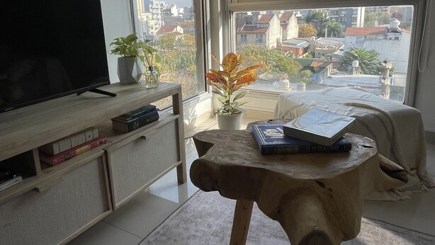 A modern living room with a flat-screen TV, a wooden coffee table with books, a small potted plant, and a view of a cityscape through large windows. - MORON GRAN ATARDECER APARTMENT (Morón)