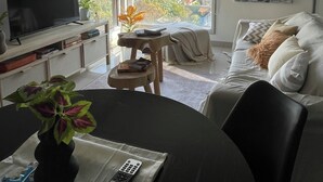 A modern living room with a flat-screen TV, a dining table with a vase of flowers, a sofa with cushions, and a large window offering a city view. - MORON GRAN ATARDECER APARTMENT (Morón)