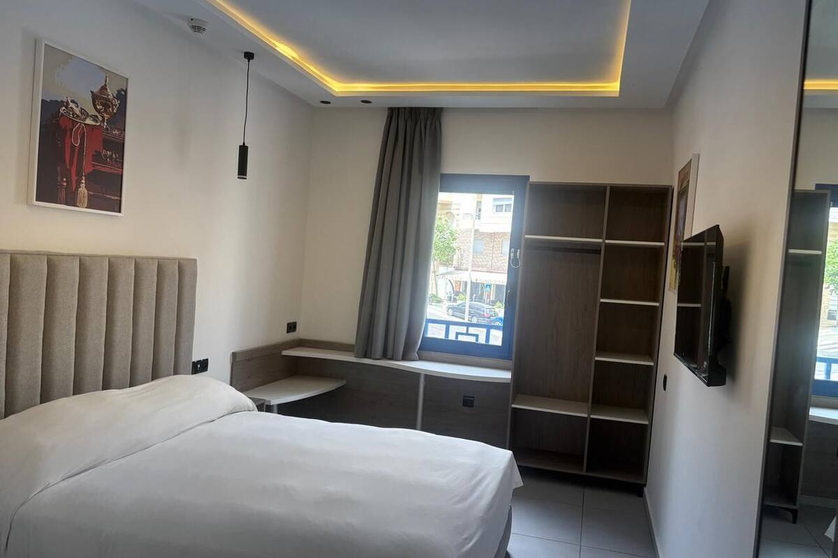A modern hotel room with a bed, a TV, a window with curtains, and a built-in wardrobe.