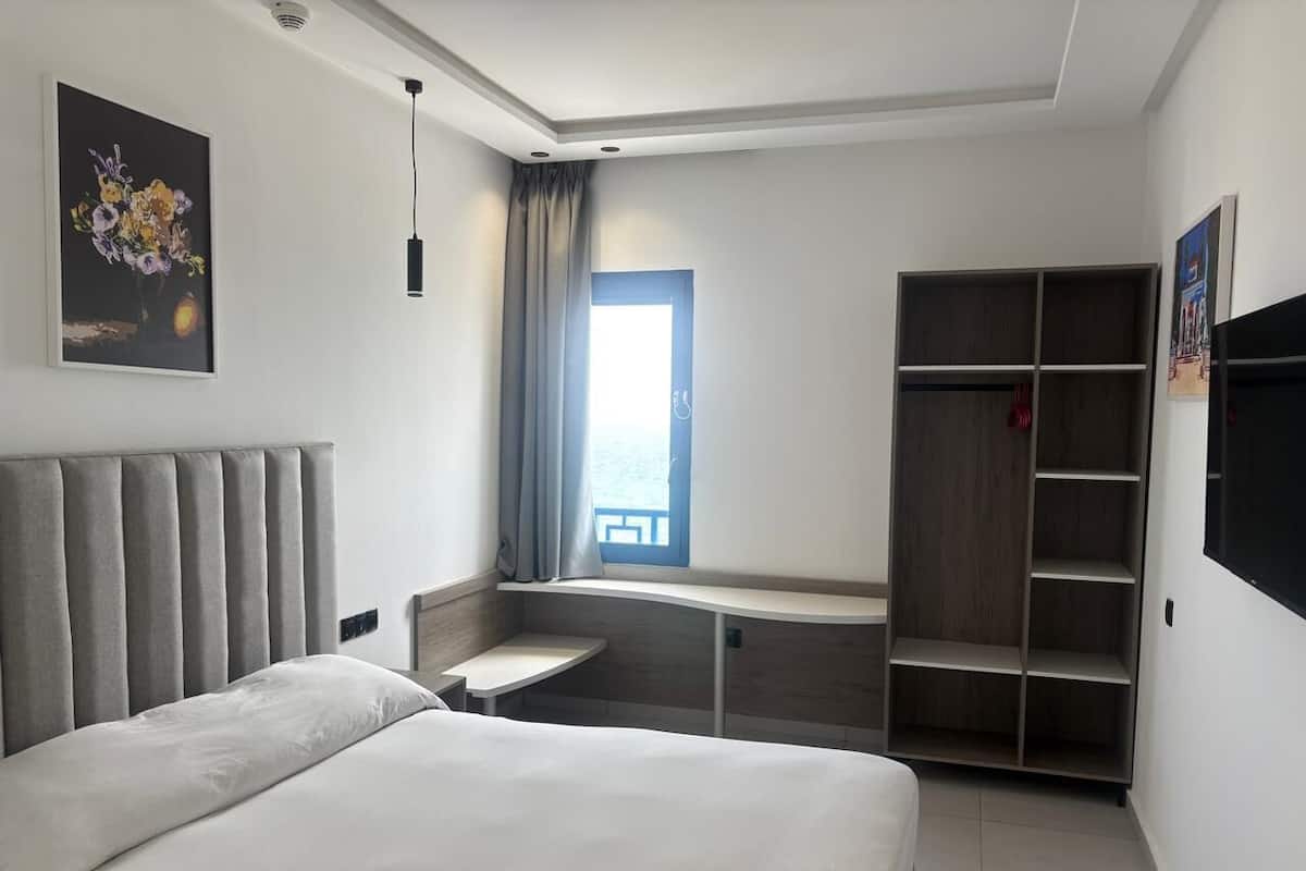 A modern hotel room with a large bed, a desk, a wardrobe, and a window with a view.