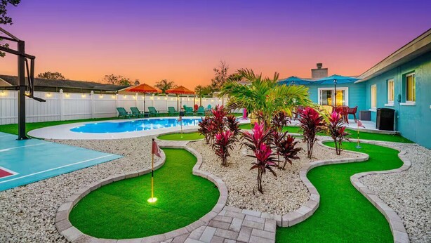 A pool area with a putting green, surrounded by palm plants and a white fence.