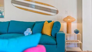 A teal sofa with yellow and blue cushions, a woven lamp, and a surfboard decoration on the wall.
