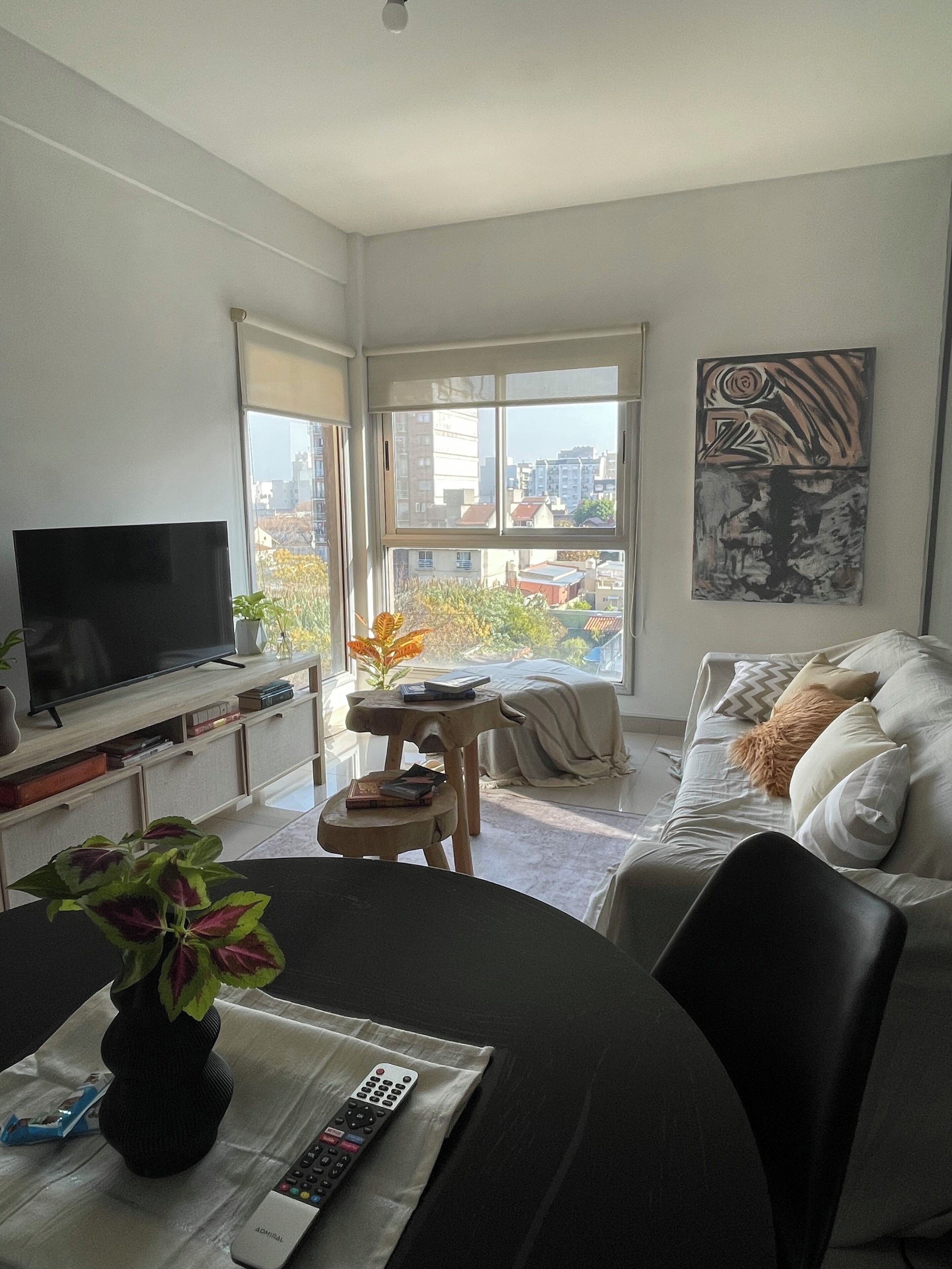 Uma sala de estar moderna com uma televisão de tela plana, uma mesa de jantar com um vaso de flores, um sofá com almofadas e uma grande janela com vista para a cidade.