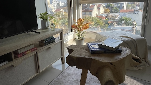 A modern living room with a flat-screen TV, a wooden coffee table with books, a small potted plant, and a view of the city through large windows. - MORON GREAT SUNSET (Morón)