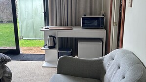 Een compacte leefruimte met een grijze bank, een klein wit bureau met een magnetron, en een schuifdeur naar een achtertuin.