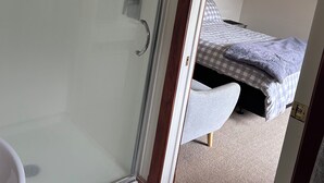 Een douche met glazen wanden, een handdoek hangt aan de deur, een bed met een geruit beddegoed en een vloerbedekking.