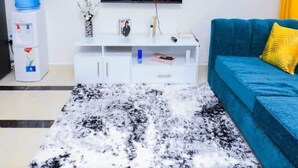 A living room with a flat-screen TV displaying a fire, a blue sofa with yellow pillows, a white entertainment unit, and a black and white rug. - Triad Apartment (Nairobi)