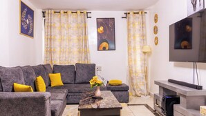 A living room with a grey sectional sofa, a coffee table with a vase of yellow flowers, and a flat-screen TV on a stand. - Triad Apartment (Nairobi)