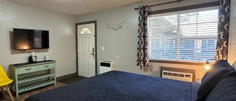 Un dormitorio con cama, televisor, ventana con cortinas, puerta y una mesita de noche con lámpara.