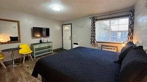 Une chambre avec un grand lit, un téléviseur fixé au mur, un bureau avec une chaise et une fenêtre avec des stores.