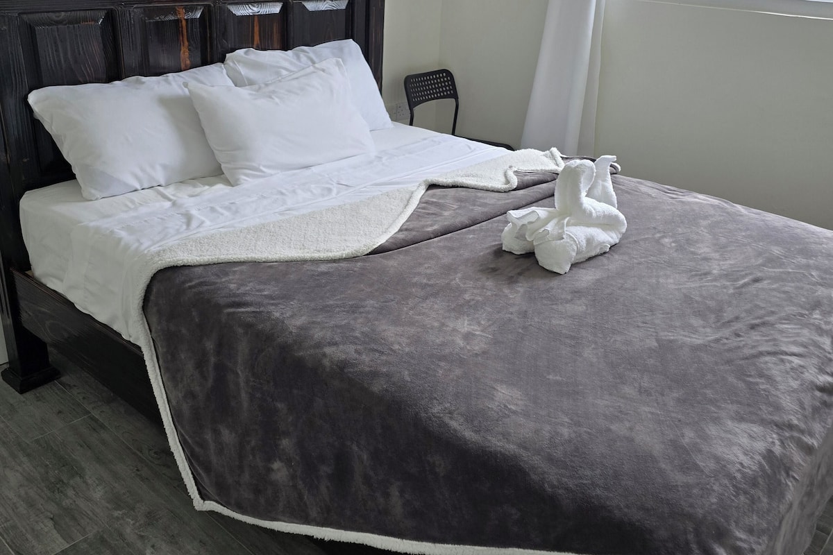 Un lit bien fait, avec du linge de lit blanc et une couverture grise, une chaise noire et une fenêtre donnant sur l’ext érieur.