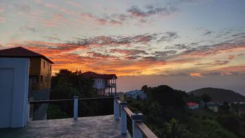 A residential area with houses and a balcony overlooking a hilly landscape during sunset.