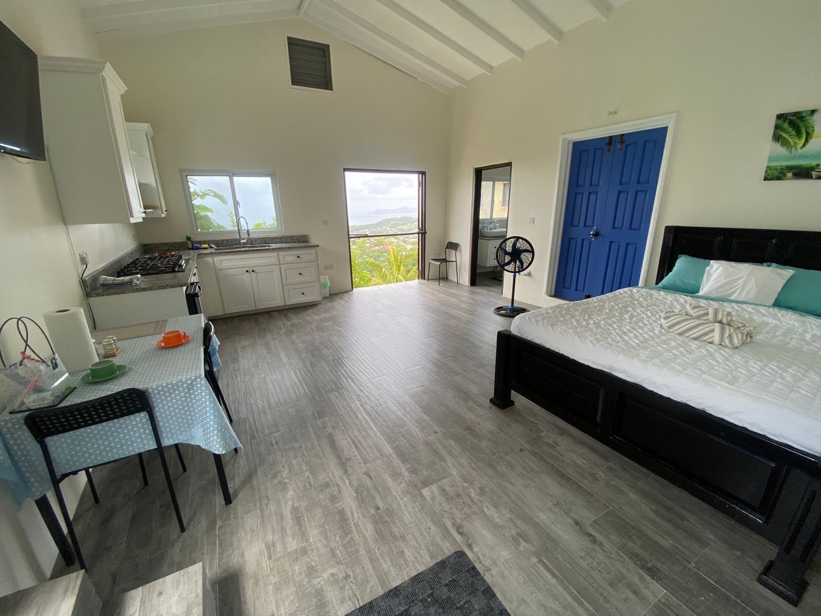 A bedroom with a bed, a desk with a chair, a kitchenette, and a view of the outdoors.