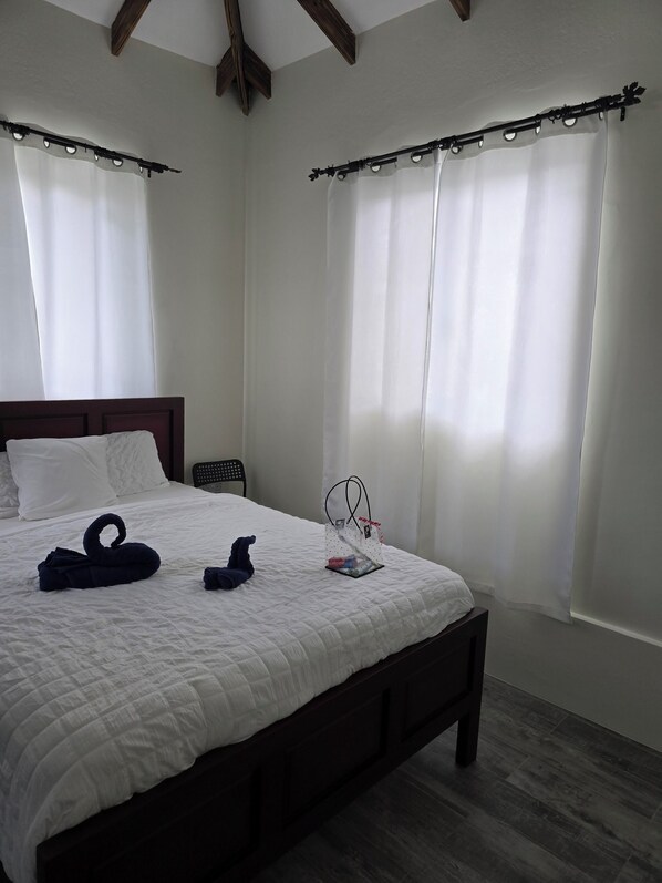 A neatly made bed with a white quilt, a wooden headboard, and a small bedside table with a clear plastic bag containing toiletries. - WindBlow Villa (Belmont)