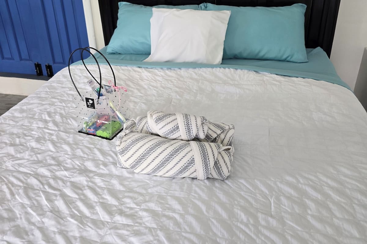 Un lit avec une couette blanche, deux oreillers (un blanc, un turquoise) et une couverture à rayures. Il y a une petite table avec une bouteille et une tasse. Un tableau représentant un bateau est accroché au mur.