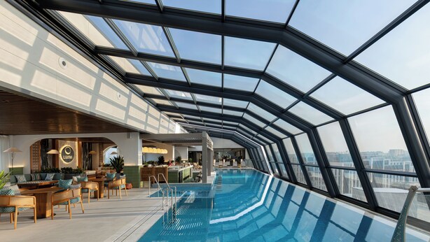 A modern indoor swimming pool with a glass ceiling, seating area, and a bar. - Doubletree By Hilton Tashkent (Tashkent)