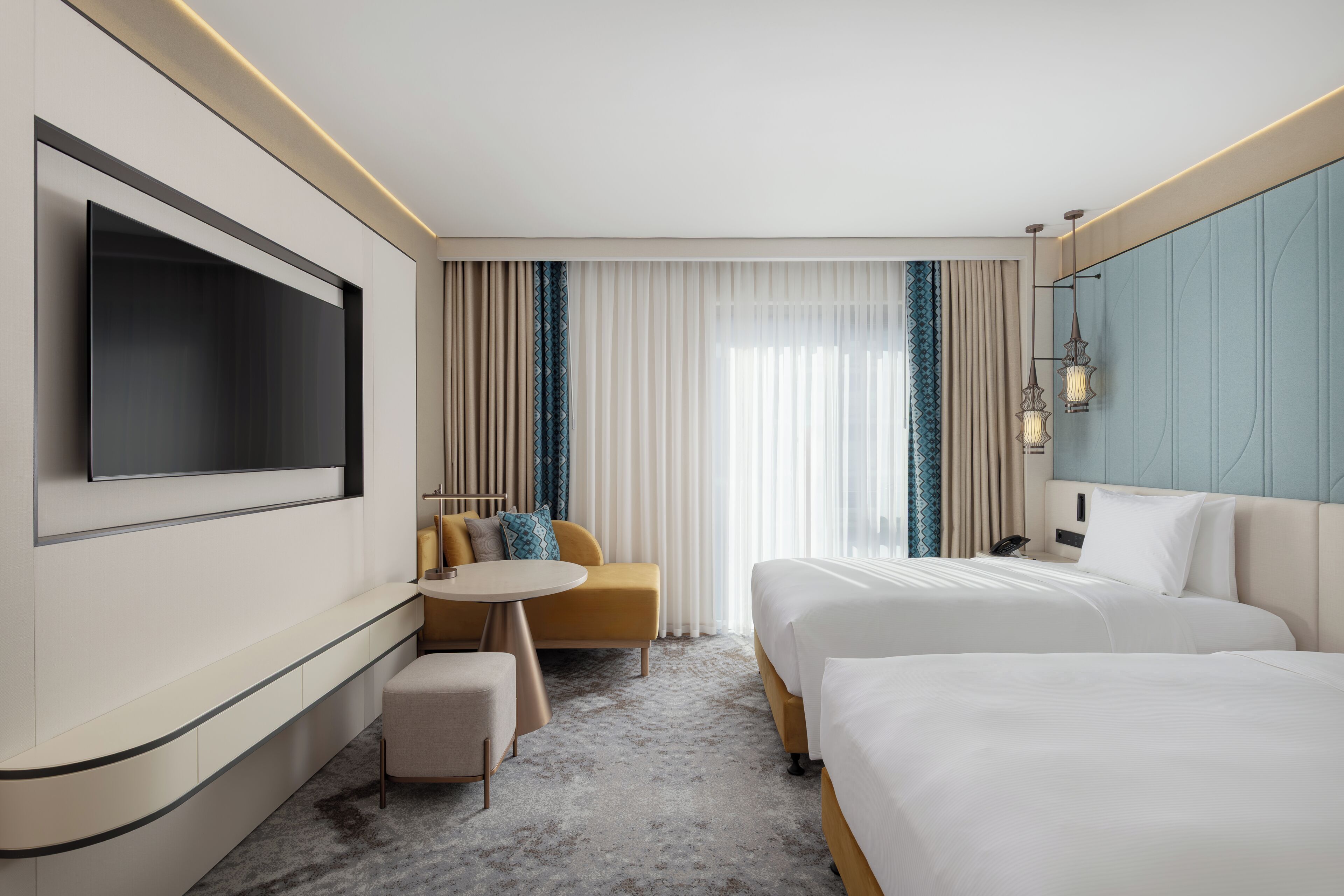 A hotel room with a TV, two beds, a sofa, a small table, and a large window with curtains.