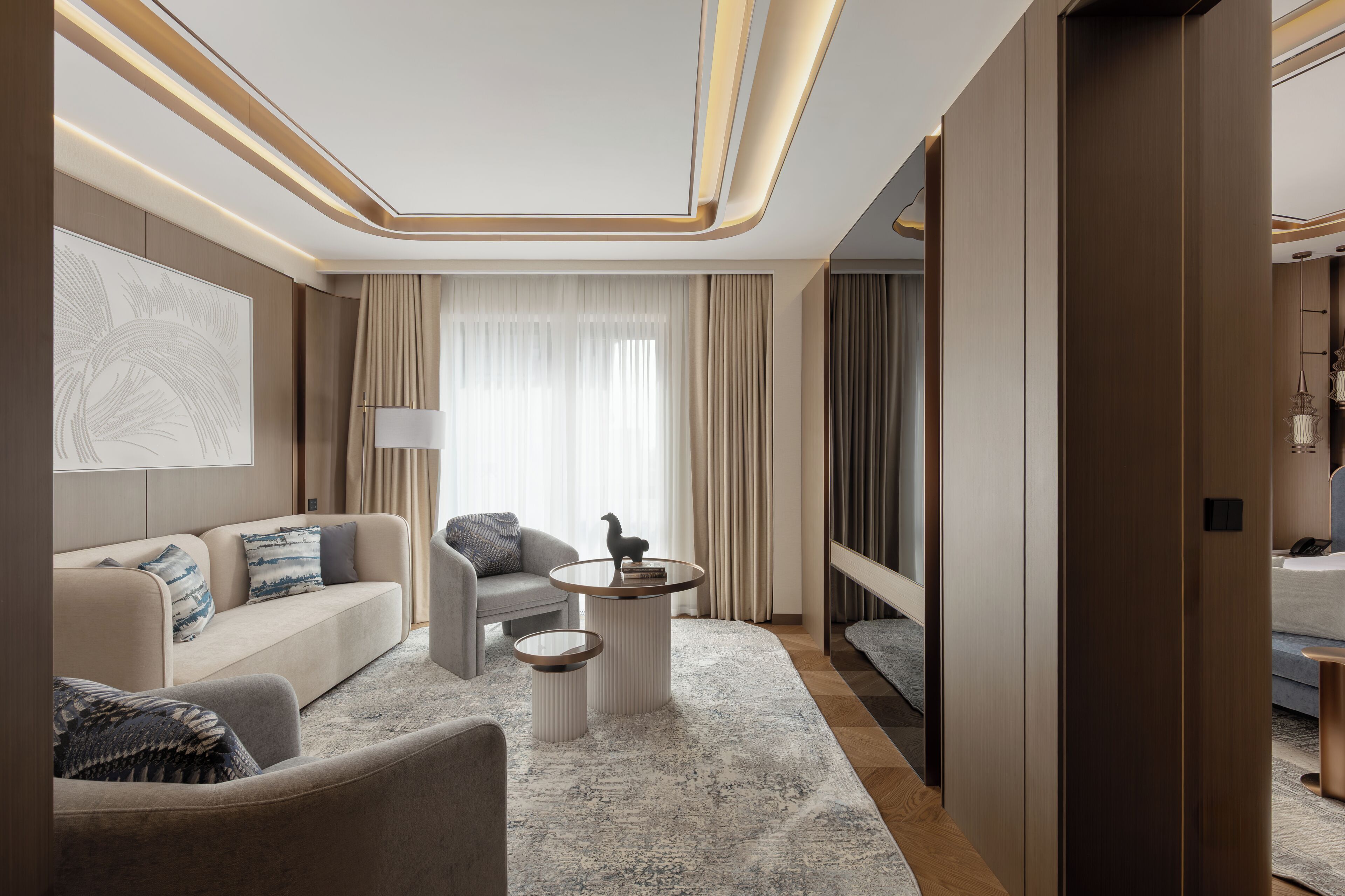 A modern hotel room with a sofa, armchair, two side tables, a lamp, and a large window with curtains.