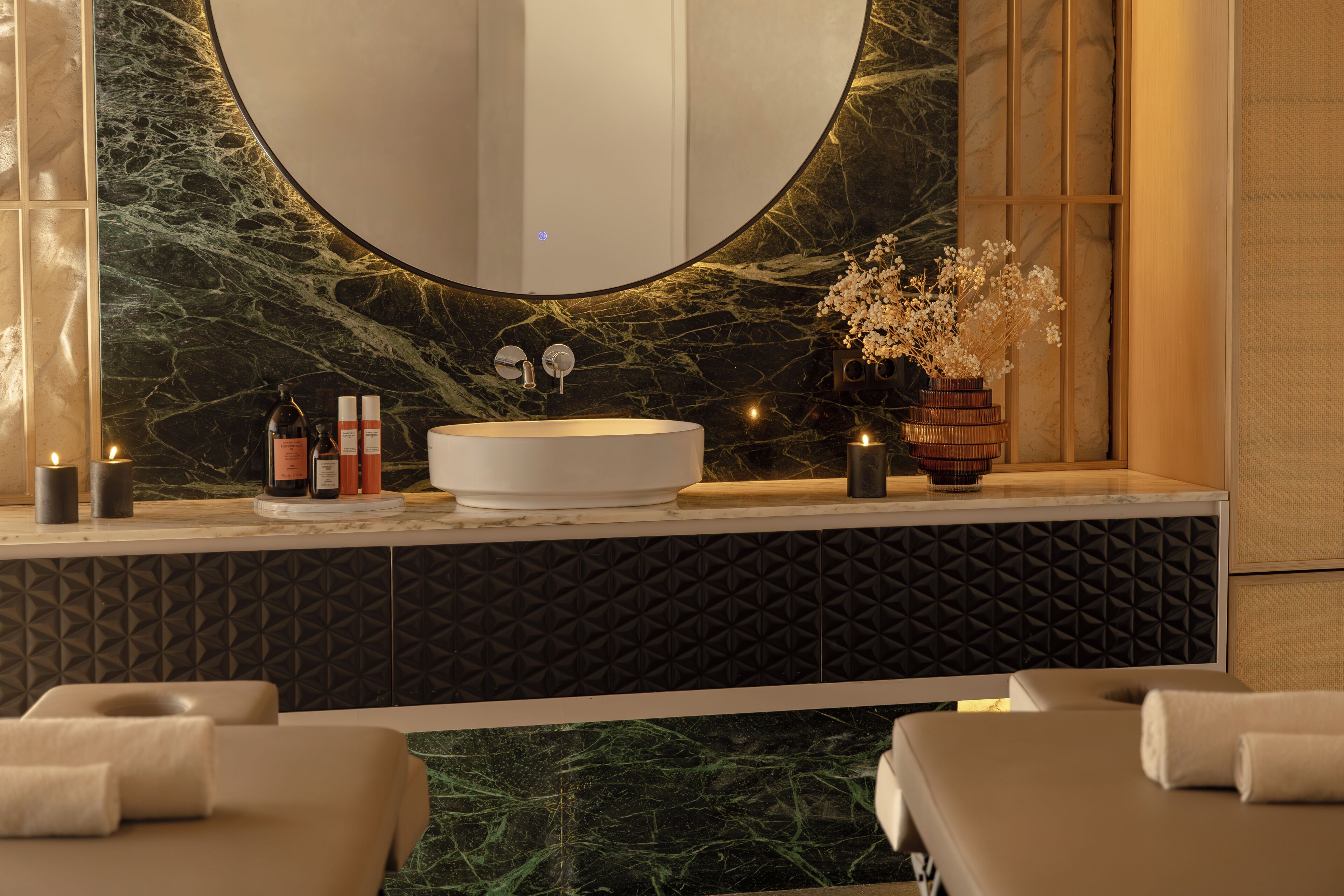 A bathroom with a round mirror, a sink, candles, and a vase with flowers.
