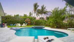 A swimming pool with a hot tub, surrounded by palm trees and a wooden playhouse.