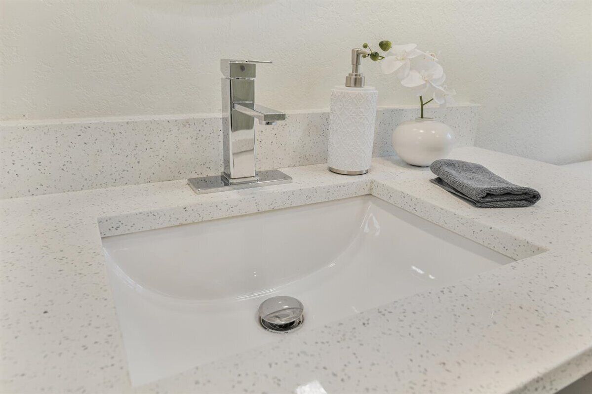 A bathroom with a white speckled countertop, a chrome faucet, a soap dispenser, a vase with an orchid, and a folded towel.