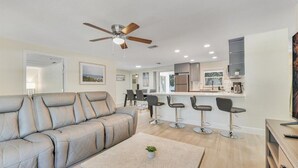 A modern living room with a beige sofa, a coffee table, and a ceiling fan. There is a kitchen area with bar stools and a refrigerator in the background.