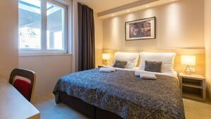Een hotelkamer met een groot bed, een bureau en een stoel. Er is een raam met gordijnen, een aan de muur bevestigd schilderij en een nachtkastje met een lamp.