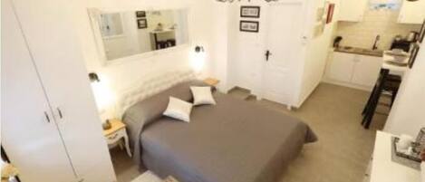 A bedroom with a large bed, a bedside table, a nightstand, a dresser, a mirror, a chandelier, and a kitchenette with a sink and stove.
