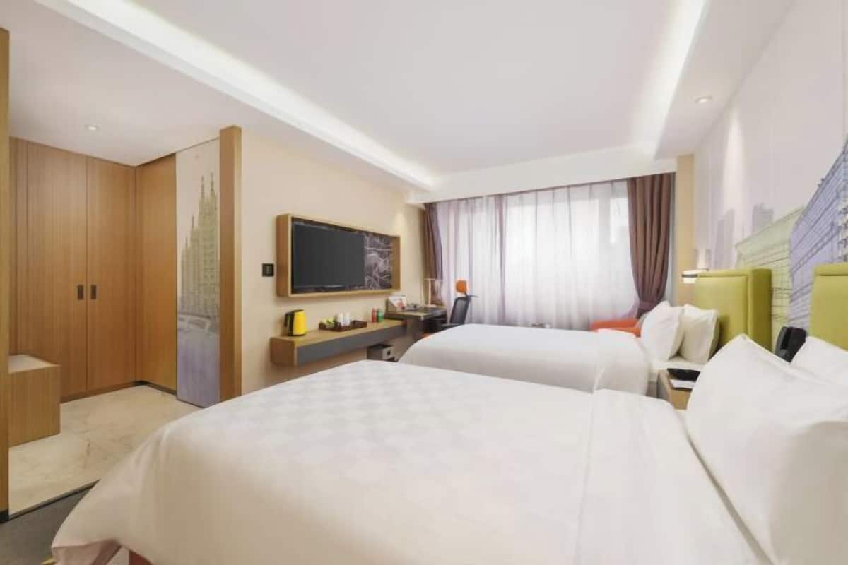 A hotel room with two beds, a desk, a television, and a large window with curtains.