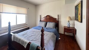 Un dormitorio con cama de madera, ventana con persianas, mesita de noche con lámpara y un cuadro en la pared.