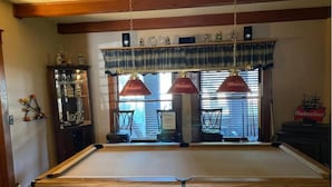 A pool table with a wooden frame, two red pendant lamps, and a window with blinds.