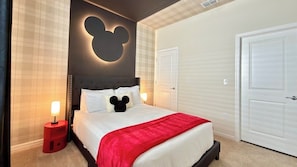 Quarto de hotel com uma cama, luminária em formato de cabeça do Mickey Mouse, parede de destaque vermelha e porta branca.