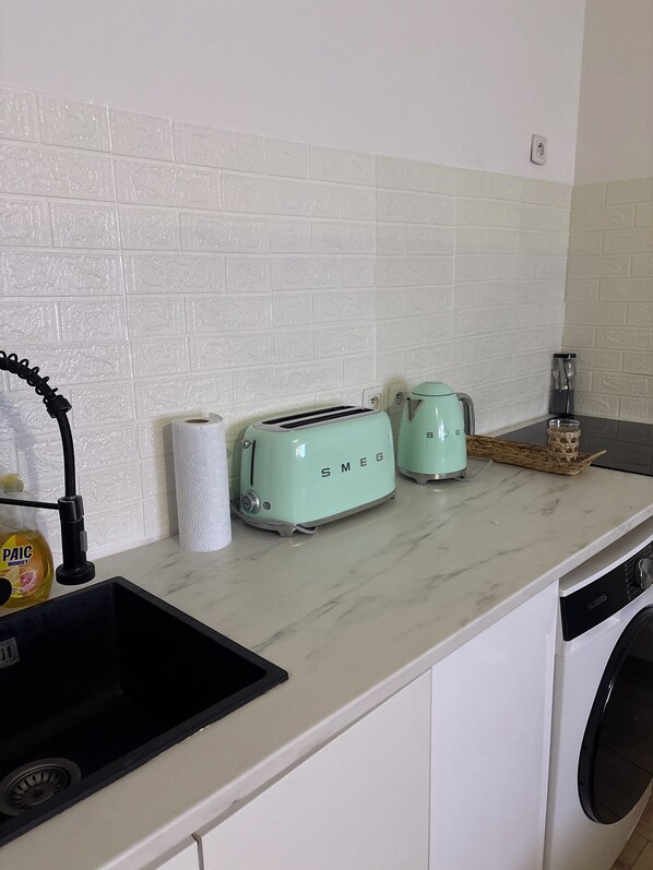 Una cocina con azulejos blancos tipo metro, un lavamanos, un grifo negro, dos tostadoras Smeg, una cafetera y una lavadora.