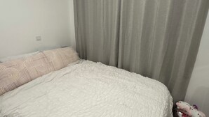 Une chambre à coucher avec un lit, des rideaux gris, une couvre-lit blanche à motifs et une petite poubelle contenant un jouet en peluche.