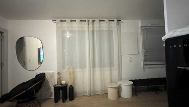 Une pièce avec un plancher en bois, une chaise noire, un miroir rond, un rideau blanc et un meuble noir.