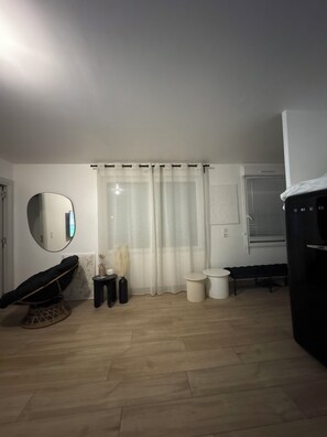 Una habitación con piso de madera, una silla negra, un espejo redondo, una cortina blanca y un mueble negro.