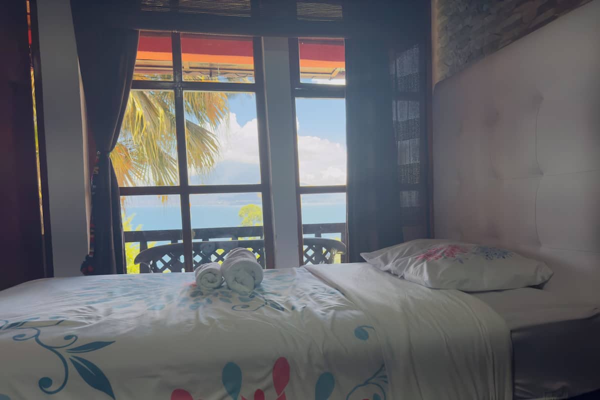 A room with a bed, a view of the sea, and a balcony with a wooden railing.