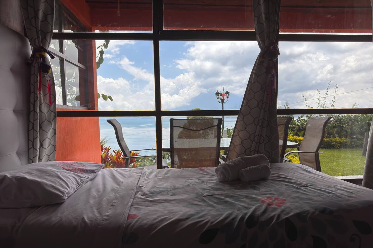 A bedroom with a large window, a bed with a white comforter, and a view of the sky and greenery outside.